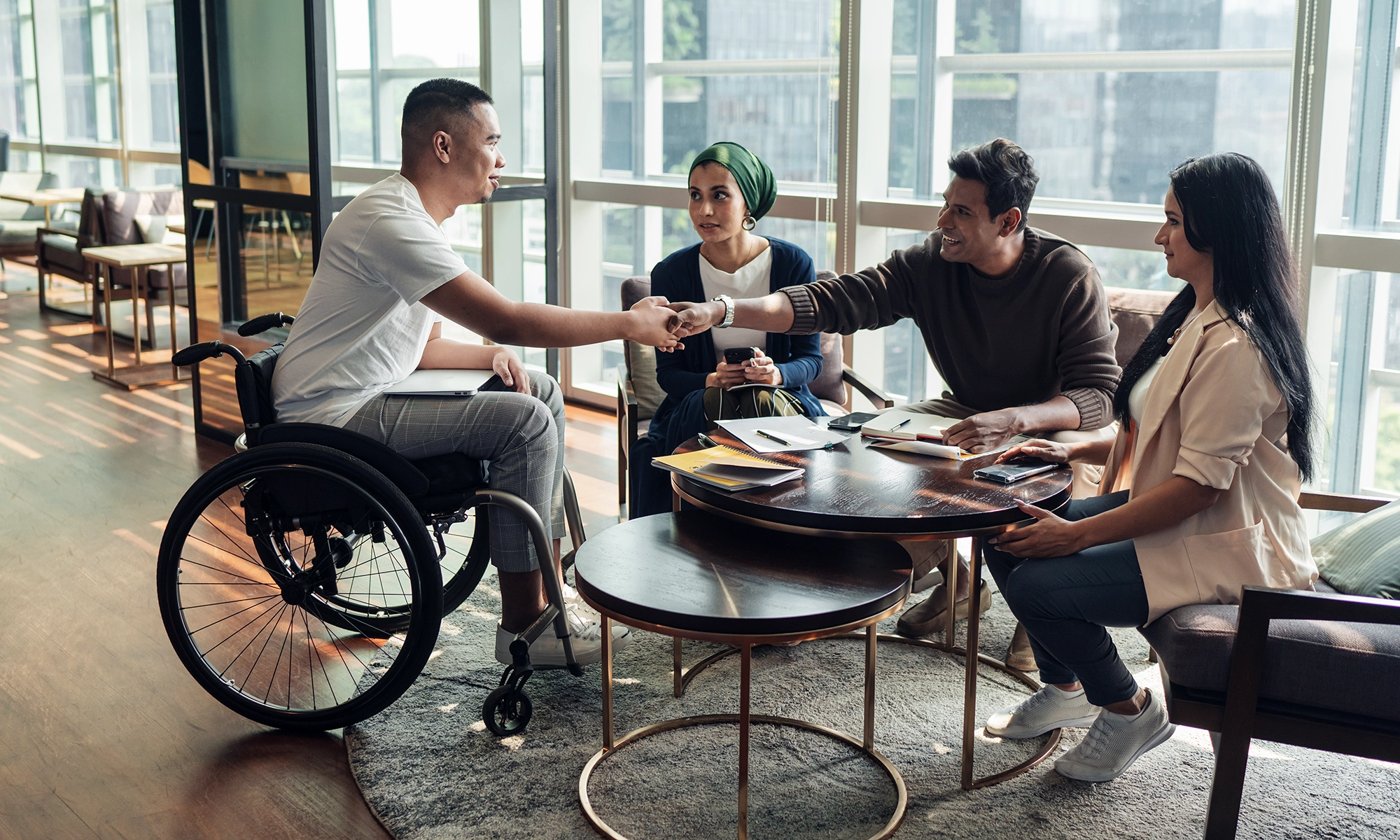 Man in a wheelchair shakes hand with a man at a table. There are two other people sitting at the table watching.