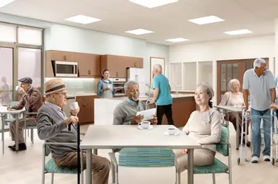 Seniors sit around a square table in the wellness kitchen, while a staff member provides meals from behind a counter