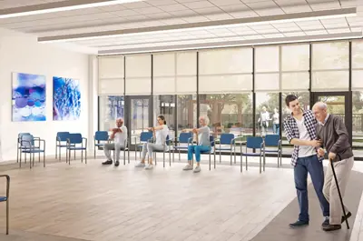 A large open area with windows overlooking an outdoor social area. Chairs are lined up for residents to use for light physical activity. A male employee helps an elderly man with a can walk through the space