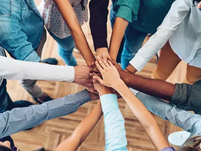 Close up of a group of diverse people in a circle each put one hand in towards the centre to indicate working together
