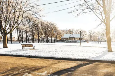 The sun shines over a snow-covered park. A lone bench is empty