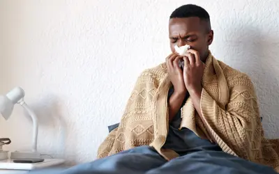 Man blowing nose into tissue while sitting in bed with a blanket