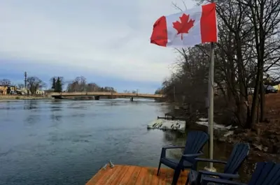 A Canadian flag flies from a pole on a deck overlooking a lake. There are three Muskoka chairs on the deck.