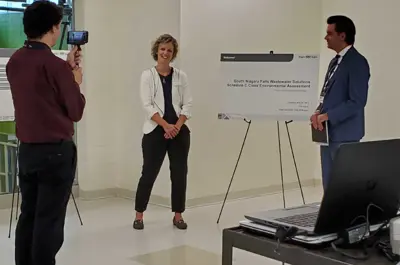 A woman stands by an information board while a man films her. Another man looks on from the side